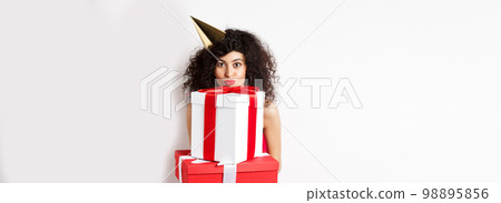 Cute birthday girl with curly hair and party hat, holding gifts and looking happy at camera, standing against white background Cute birthday girl with curly hair and party hat, holding gifts and looking happy at camera, standing against white background 98895856