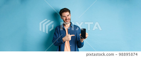 Smiling caucasian man showing on empty smartphone screen, pointing at mobile phone and looking satisfied, standing on blue background 98895874