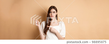 Hesitant young woman look unsure, pointing fingers right while making decision, frowning with puzzled face, standing on beige background 98895875