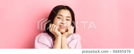 Close-up portrait of dreamy and romantic asian girl, lean face on hands and smiling, looking with admiration and happiness, standing against pink background 98895876