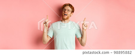 Amazed redhead guy in glasses checking out special deal, looking and pointing fingers up with amazed face, standing over pink background 98895882