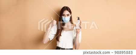 Covid-19 and preventive measures concept. Smiling girl in medical mask show thumb up and hand sanitizer, recommend product for disinfection, beige background 98895883
