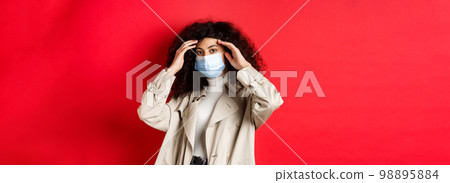 Covid-19, pandemic and quarantine concept. Stylish young woman with curly hair, going outside in medical mask and trench coat, fixing haircut, standing on red background Covid-19, pandemic and quarantine concept. Stylish young woman with curly hair, going outside in medical mask and trench coat, fixing haircut, standing on red background 98895884