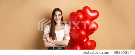 Cheerful young woman looking happy on Valentines day, standing near hearts balloons with arms crossed, smiling at camera, standing on beige background Cheerful young woman looking happy on Valentines day, standing near hearts balloons with arms crossed, smiling at camera, standing on beige background 98895894