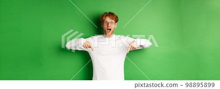 Surprised and excited young redhead man scream of joy and amazement, checking out awesome promo, pointing fingers down, standing over green background Surprised and excited young redhead man scream of joy and amazement, checking out awesome promo, pointing fingers down, standing over green background 98895899