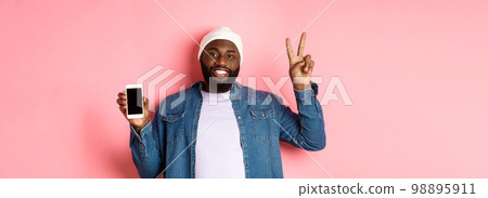 Online shopping and technology concept. Happy Black guy in beanie and denim shirt showing mobile screen and peace sign, standing over pink background Online shopping and technology concept. Happy Black guy in beanie and denim shirt showing mobile screen and peace sign, standing over pink background 98895911