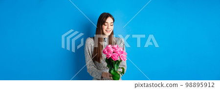 Valentines day concept. Romantic girl in dress looking happy at flowers, smiling at bouquet of pink roses, standing on blue background Valentines day concept. Romantic girl in dress looking happy at flowers, smiling at bouquet of pink roses, standing on blue background 98895912
