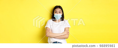 Covid, health care and pandemic concept. Asian woman in white t-shirt and medical mask cross arms on chest and looking surprised at camera Covid, health care and pandemic concept. Asian woman in white t-shirt and medical mask cross arms on chest and looking surprised at camera 98895981