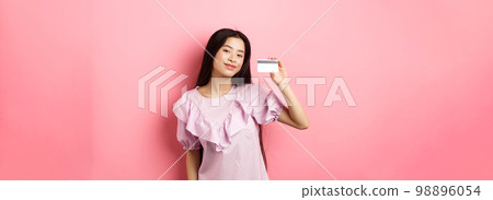 Shopping. Young korean girl in dress showing credit card, smiling satisfied at camera, standing on pink background Shopping. Young korean girl in dress showing credit card, smiling satisfied at camera, standing on pink background 98896054