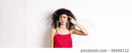 Shocked and confused caucasian woman in red dress scolding someone crazy, pointing finger at head and frowning, standing over white background 98896112