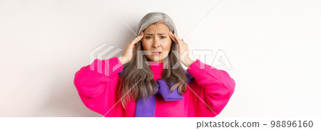 Close-up of fashionable asian senior woman feeling unwell, touching head and grimacing from headache, complain on migraine, white background Close-up of fashionable asian senior woman feeling unwell, touching head and grimacing from headache, complain on migraine, white background 98896160
