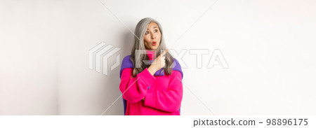 Curious asian senior female checking out promo, looking and pointing finger left, standing over white background 98896175