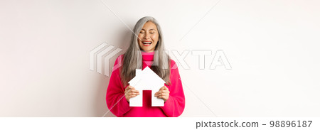 Real estate. Happy asian grandmother laughing with eyes closed, holding paper house model, dreaming about paper house model, standing over white background 98896187