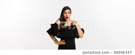 Fashion and beauty. Shocked woman pointing at herself and looking at camera, being accused, standing in black dress over white background Fashion and beauty. Shocked woman pointing at herself and looking at camera, being accused, standing in black dress over white background 98896347