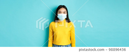 Covid-19, social distancing and pandemic concept. Teenage asian woman in medical mask, looking at camera and standing against blue background in yellow sweater Covid-19, social distancing and pandemic concept. Teenage asian woman in medical mask, looking at camera and standing against blue background in yellow sweater 98896436
