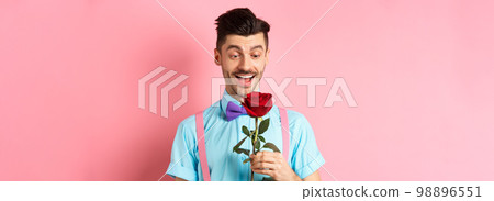 Valentines day and romance concept. Happy boyfriend looking at red rose while waiting for lover in bow-tie, standing on pink romantic background 98896551