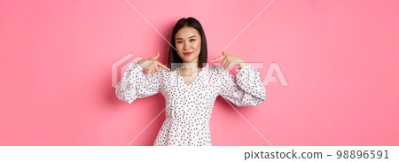 Beautiful asian woman looking confident, pointing at herself and smiling, showing logo on chest, standing over pink background 98896591