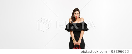 Beauty and fashion concept. Full length if silly young woman pouting and looking surprised, holding purse, wearing heels and black dress, white background 98896633