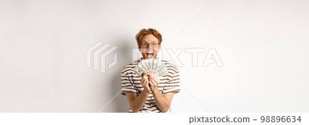 Lucky young man with red hair showing dollars, winning money and screaming of happiness, holding prize cash, standing over white background 98896634
