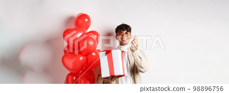 Valentines day and romance concept. Handsome young man showing surprise gift for lover and finger heart, smiling, standing near festive red balloons, white background 98896756