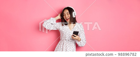 Carefree asian woman having fun, dancing and listening music in headphones, holding mobile phone, standing in dress over pink background 98896782