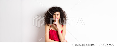 Angry elegant woman in red dress hushing and frowning, tell to be quiet, asking for silence, standing over white background 98896790