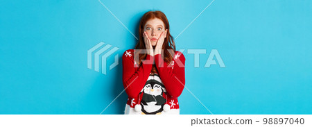 Winter holidays and Christmas Eve concept. Impressed and speechless redhead girl gasping, staring at camera with disbelief, standing in xmas sweater against blue background 98897040