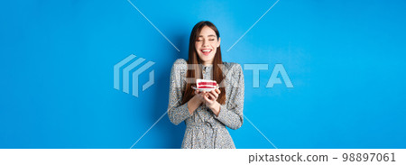 Dreamy birthday girl making wish with closed eyes and happy smile, blowing cake candle, standing on blue background Dreamy birthday girl making wish with closed eyes and happy smile, blowing cake candle, standing on blue background 98897061