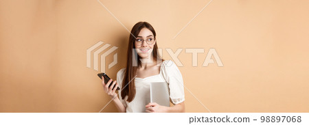 Image of young stylish woman going on work, holding laptop and smartphone, looking aside at empty space, standing on beige background Image of young stylish woman going on work, holding laptop and smartphone, looking aside at empty space, standing on beige background 98897068