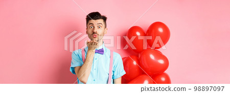 Valentines day concept. Attractive and funny man in bow-tie, blowing air kiss to lover, standing near pink background and red hearts balloons 98897097