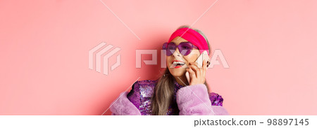 Fashion. Close-up of fashionable asian senior woman in sunglasses and disco outfit talking on phone, laughing and smiling, standing on pink background 98897145