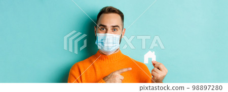 Real estate and coronavirus pandemic concept. Close-up of adult man in medical mask pointing at small paper house, standing over light blue background Real estate and coronavirus pandemic concept. Close-up of adult man in medical mask pointing at small paper house, standing over light blue background 98897280