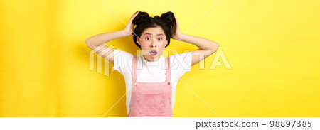 Shocked asian girl gasping worried, checking her hairstyle, standing anxious on yellow background 98897385