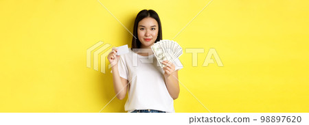 Beautiful asian woman with short dark hair, wearing white t-shirt, showing money in dollars and plastic credit card, standing over yellow background 98897620