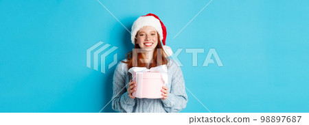 Winter holidays and Christmas Eve concept. Smiling redhead girl in sweater and Santa hat, holding New Year gift and looking at camera, standing against blue background 98897687