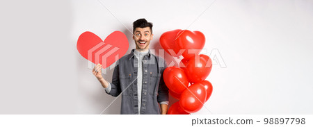 Valentines day and love concept. Cheerful funny guy showing heart cutout, standing near romantic balloons and smiling excited at camera, white background Valentines day and love concept. Cheerful funny guy showing heart cutout, standing near romantic balloons and smiling excited at camera, white background 98897798