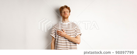 Young bearded redhead man holding hand on heart and looking patriotic, standing during national anthem, standing over white background Young bearded redhead man holding hand on heart and looking patriotic, standing during national anthem, standing over white background 98897799