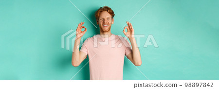 Attractive young man in t-shirt smiling satisfied, nod in approval and showing OK sign, approve and agree with something cool, standing over turquoise background Attractive young man in t-shirt smiling satisfied, nod in approval and showing OK sign, approve and agree with something cool, standing over turquoise background 98897842