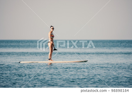 Sea woman sup. Silhouette of happy middle aged tanned woman in rainbow bikini, surfing on SUP board, confident paddling through water surface. Idyllic sunset. Active lifestyle at sea or river. 98898184