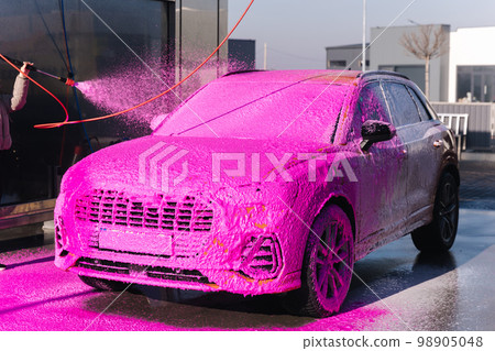 Young woman cleaning her car on self service car wash. Female spread soap using jet sprayer. Pink foam on the car Young woman cleaning her car on self service car wash. Female spread soap using jet sprayer. Pink foam on the car 98905048