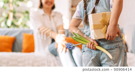 A child holding a gift and flowers behind her back in front of her mother, mother's day A child holding a gift and flowers behind her back in front of her mother, mother's day 98906414