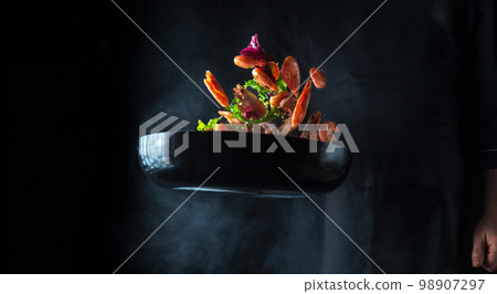 Chef cooks shrimp in a pan with vegetables. Cooking seafood, healthy vegetarian food and food on a black background. Freezing in motion. Free advertising space Chef cooks shrimp in a pan with vegetables. Cooking seafood, healthy vegetarian food and food on a black background. Freezing in motion. Free advertising space 98907297