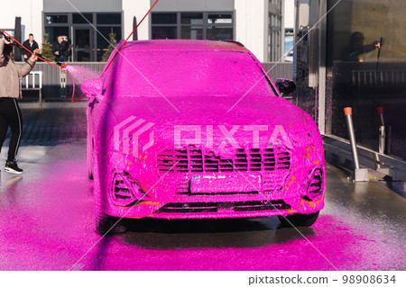 Young woman cleaning her car on self service car wash. Female spread soap using jet sprayer. Pink foam on the car Young woman cleaning her car on self service car wash. Female spread soap using jet sprayer. Pink foam on the car 98908634