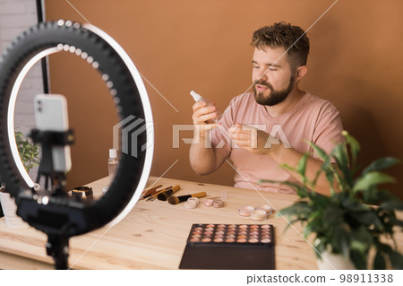 Famous blogger. Cheerful male vlogger showing cosmetics products while recording video and giving advices for his beauty blog. Make-up artist and recording beauty vlog concept Famous blogger. Cheerful male vlogger showing cosmetics products while recording video and giving advices for his beauty blog. Make-up artist and recording beauty vlog concept 98911338