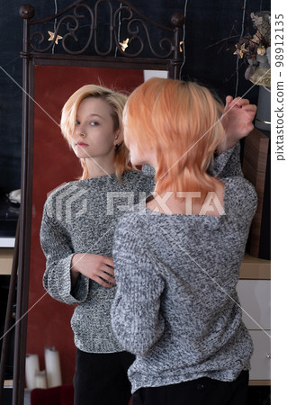 teenager girl looks in the mirror indoor, carefully examines her hairstyle teenager girl looks in the mirror indoor, carefully examines her hairstyle 98912135