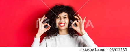 Close-up portrait of smiling woman with curly hair and red lips, showing okay gesture and looking satisfied, praise good product, standing on red background 98913785