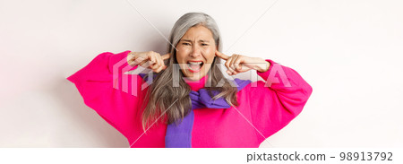 Close-up of annoyed and bothered asian senior woman screaming with shut ears, disturbed by loud noise, standing over white background Close-up of annoyed and bothered asian senior woman screaming with shut ears, disturbed by loud noise, standing over white background 98913792