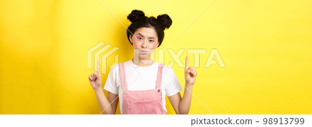 Annoyed and tired asian girl with pink glamour makeup, pointing fingers up and look bothered, standing on yellow background 98913799