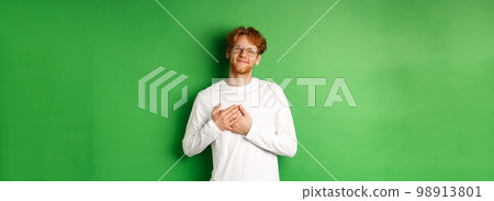 Handsome young man in glasses and white long-sleeve smiling, holding hands on heart with grateful face, thanking you, standing over green background 98913801