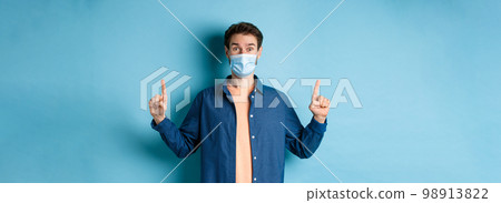 Happy guy in medical mask feeling excited about good news, pointing fingers up and smiling, standing on blue background 98913822